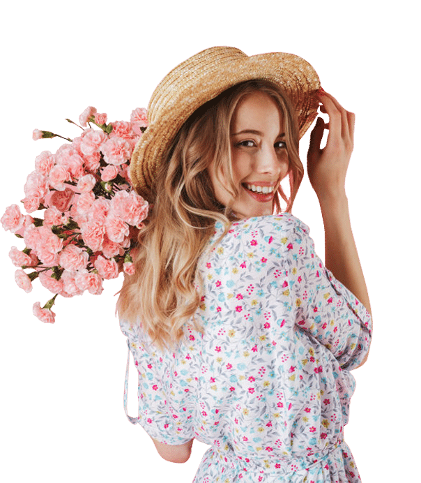 Smiling woman in floral dress and straw hat holding pink flowers.