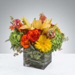 Bright floral arrangement with yellow lilies, orange roses, and green foliage in a glass cube vase.