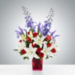A vibrant patriotic-themed flower arrangement in a translucent red glass vase, featuring white lilies, red roses, and tall blue delphinium and iris accents.