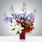 A vibrant floral arrangement featuring white lilies, purple irises, and red roses in a square pink glass vase, set against a neutral gradient background