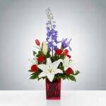 A patriotic-themed floral arrangement in a vibrant red glass vase featuring white lilies, red roses, red tulips, and tall blue delphinium accents.