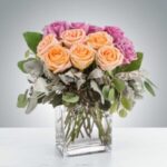 A bouquet of peach roses and purple flowers in a clear square vase.