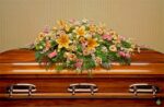 A vibrant floral spray consisting of lilies, carnations, and greenery arranged on top of a polished wooden casket.