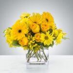 Bright yellow flowers arranged in a clear square vase.