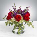 Elegant bouquet of red roses with purple flowers and greenery in a clear glass vase.