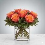 A petite floral arrangement featuring vibrant coral and orange roses nestled in green variegated foliage, presented in a clear square glass vase on a white marble surface.
