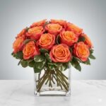 A compact arrangement of vibrant orange and coral roses with bi-color petals, accented with variegated green and white leaves, presented in a clear square glass vase on a marble surface.