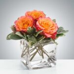 A petite floral arrangement featuring three vibrant orange and pink bi-color roses nestled among lush green and variegated foliage, presented in a modern clear glass cube vase filled with water.
