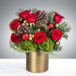 Festive floral arrangement with red roses, pine cones, and greenery in a gold vase.