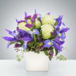 A vibrant floral arrangement in a square white ceramic vase featuring purple irises with yellow centers, creamy white roses, and small clusters of purple statice flowers.