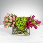 A lush floral arrangement in a rectangular glass vase featuring a central cluster of vibrant green hydrangeas flanked by light pink and deep magenta tulips on a white marble surface.