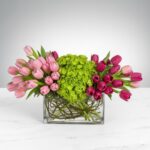 A horizontal floral arrangement in a rectangular glass vase featuring light pink tulips on the left, a large green hydrangea in the center, and dark pink/magenta tulips on the right.