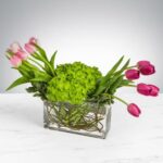 A horizontal floral arrangement in a rectangular clear glass vase featuring a cluster of lime green hydrangeas in the center, flanked by light pink tulips on the left and deep magenta tulips on the right, all set against a white marble surface and a soft grey gradient background.