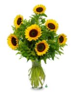 A vibrant bouquet of seven large yellow sunflowers with dark brown centers, arranged with lush green filler foliage in a clear glass vase against a solid white background.