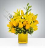 A lush, monochromatic yellow floral arrangement featuring roses, lilies, and tulips in a translucent yellow glass square vase, accented with green hypericum berries and eucalyptus.