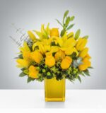 A vibrant premium bouquet of yellow roses and lilies arranged in a square yellow glass vase with green berries and eucalyptus accents.