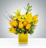 A vibrant all-yellow floral arrangement featuring roses, lilies, and tulips in a translucent yellow glass cube vase, accented with green hypericum berries and eucalyptus.