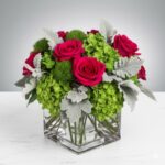 A vibrant floral arrangement featuring hot pink roses, green hydrangeas, green trick dianthus, and silvery dusty miller leaves in a square clear glass vase.