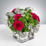 A vibrant flower arrangement featuring deep pink roses, green hydrangea, and silvery Dusty Miller foliage in a square glass vase.