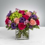 A vibrant and dense floral arrangement in a square glass vase featuring hot pink roses, peach roses, purple lisianthus, green hydrangea, and purple freesia against a neutral grey and white background.