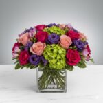 A lush, rounded floral arrangement in a square glass vase featuring vibrant pink roses, peach roses, purple lisianthus, and lime green hydrangeas on a white marble surface.