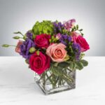 A vibrant spring flower bouquet in a square glass vase featuring hot pink roses, light peach roses, purple lisianthus, and green hydrangea with small purple statice accents.