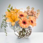 A vibrant bouquet of orange lilies and gerbera daisies in a round glass vase.