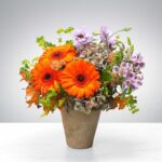 Bright orange gerbera daisies and delicate purple flowers in a rustic pot.