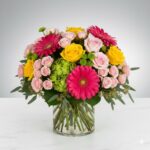 A vibrant and dense floral arrangement in a clear glass cylinder vase, featuring hot pink gerbera daisies, yellow roses, light pink spray roses, and green hydrangea, set against a neutral grey gradient background.