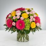A lush, colorful floral arrangement in a clear cylindrical glass vase featuring vibrant pink gerbera daisies, yellow and pale pink roses, and green hydrangea on a white marble surface.