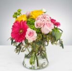 A vibrant floral arrangement in a clear glass vase featuring pink roses, a large hot pink gerbera daisy, yellow roses, and green hydrangea accents with eucalyptus leaves.