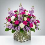 A lush and vibrant floral arrangement featuring hot pink carnations, purple stock flowers, and white carnations in a clear square glass vase.