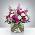 A lush floral arrangement featuring vibrant hot pink carnations, soft white carnations, and tall purple stock flowers arranged in a square glass vase filled with clear water.
