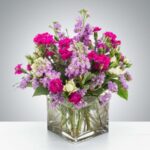 A vibrant floral arrangement featuring magenta carnations, lavender stocks, and white spray roses in a square glass vase.