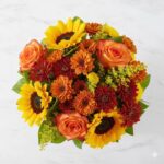 A vibrant bouquet with sunflowers, roses, and autumn leaves in a clear glass vase.