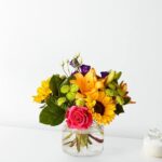 A vibrant mixed flower bouquet in a clear glass cylinder vase, featuring a large pink rose, bright yellow sunflowers, orange lilies, purple lisianthus, and green button poms, set against a clean white background.