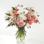 A lush and elegant floral arrangement in a clear glass vase featuring a mix of pink carnations, peach spray roses, white daisies, and pink alstroemeria with green fern accents.