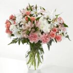 A lush floral arrangement in a clear glass vase featuring a mix of pink roses, pink carnations, white daisies, and pink alstroemeria accented with green leather leaf fern.