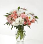 A lush floral arrangement in a clear glass vase featuring a mix of peach spray roses, white daisies, pink carnations, and light pink alstroemeria with green fern accents.