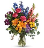A vibrant and diverse "Colors of the Rainbow" flower bouquet in a clear glass vase, featuring hot pink roses, bright orange lilies, yellow spray roses, blue delphinium, purple alstroemeria, and pink snapdragons accented with lush greenery.