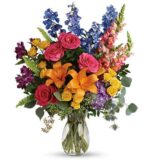 A vibrant and diverse "Colors of the Rainbow" flower bouquet in a clear glass vase, featuring hot pink roses, orange lilies, blue delphinium, yellow spray roses, and purple alstroemeria with lush greenery.