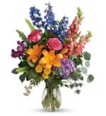 A vibrant and colorful "Colors of the Rainbow" flower bouquet in a clear glass vase, featuring blue delphinium, pink roses, orange lilies, yellow spray roses, purple hydrangea, and green snapdragons with various lush greenery.