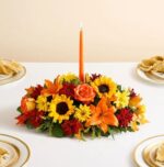A lush, horizontal autumn flower centerpiece featuring sunflowers, orange lilies, roses, and red chrysanthemums with a tall orange taper candle in the center, set on a white tablecloth with elegant place settings.