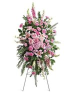 A tall, elegant standing funeral spray on a wire easel, featuring a lush arrangement of pink roses, stargazer lilies, carnations, and snapdragons with green fern accents.