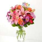 A vibrant birthday flower arrangement in a clear glass vase featuring pink lilies, orange roses, purple daisies, and a colorful "Happy Birthday" topper.