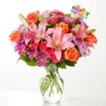 A colorful birthday flower bouquet in a clear glass vase featuring pink lilies, orange roses, and purple chrysanthemums with a "Happy Birthday" topper.