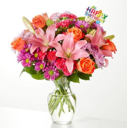 A colorful birthday flower bouquet in a clear glass vase featuring pink lilies, orange roses, and purple chrysanthemums with a "Happy Birthday" topper.