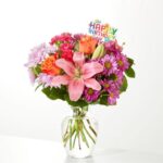 A vibrant birthday flower arrangement in a clear glass vase featuring pink lilies, orange roses, purple daisies, and a colorful "Happy Birthday" topper.
