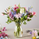 A lush pastel flower bouquet in a clear glass vase featuring pink tulips, white roses, lavender chrysanthemums, and blue larkspur, set against a neutral background with floral trimmings and scissors nearby.