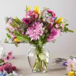 A vibrant and lush pastel flower bouquet in a clear glass vase featuring pink chrysanthemums, purple roses, and yellow tulips on a neutral tabletop.